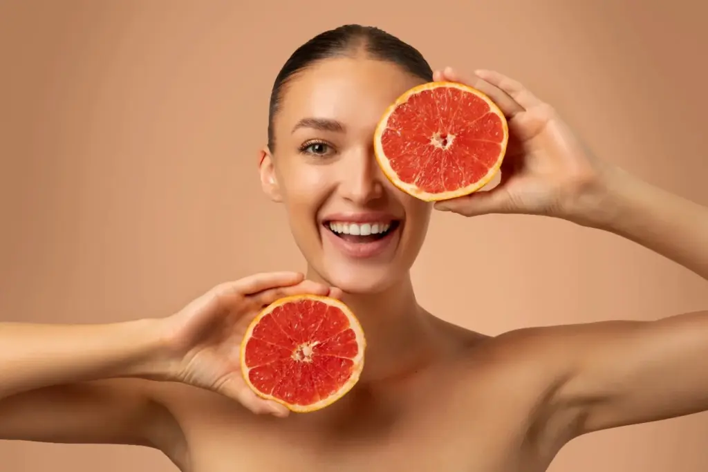 Smiling woman holding a sliced grapefruit over one eye to showcase bright Vitamin C skin benefits